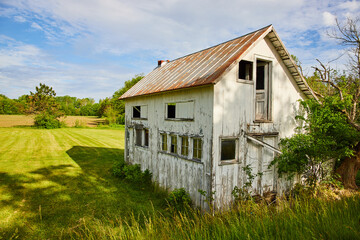 Abandoned farm house in field with busted windows and door next to dead tree