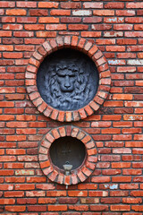 Lion head with mane and bird over fountain on tall red brick wall with circular openings to motifs