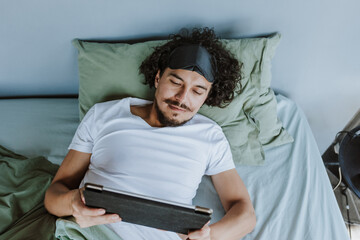 latin young man waking up and holding a tablet in bed at home in Mexico Latin America, hispanic...