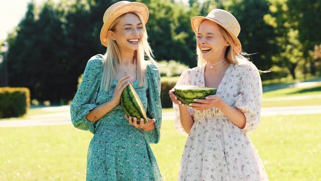 Two young beautiful smiling hipster woman in trendy summer sundress. Carefree women posing in the park in hats. Positive models having fun and hugging at sunset. They hold fresh yellow watermelon 