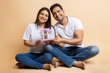 Happy young indian couple wearing casual cloths holding house toy while sitting together on floor...