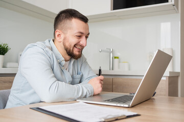 Portrait of young smiling man using laptop sitting at desk, writing in notebook. Cheerful guy browsing internet, watching webinar studying online, looking at pc screen at home.