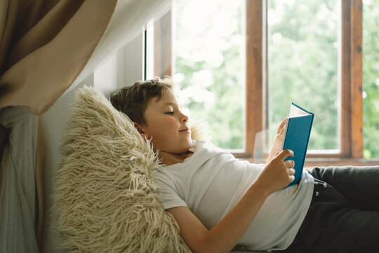 Cute boy reading a book on the windowsill near the window. Child relaxing at home on sunny day Summer. Home Schooling and education concept