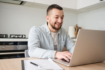 Obraz premium Portrait of young smiling man using laptop sitting at desk, writing in notebook. Cheerful guy browsing internet, watching webinar studying online, looking at pc screen at home.