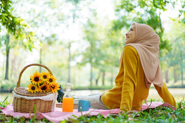 Happy Asian muslim woman sitting on the grass field in garden. Muslim woman having a picnic in garden.