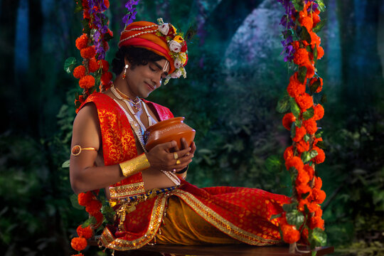 Young man dressed up as Lord Krishna and sitting on a swing with a butter pot on the occasion of Janmashtami