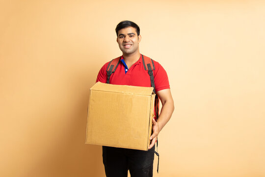 Portrait of happy Indian delivery man wearing red T-shirt holding cardboard box isolated on beige studio background, Courier boy or Parcel Service concept