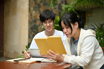 A focused Asian female college student is reading a book while her male friend is using a tablet