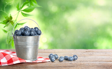 Small metal bucket with blueberries and checkered napkin on wooden table on defocus green background. Summer background