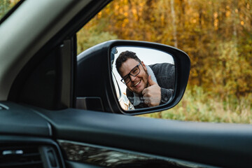 Fototapeta na wymiar A smiling man in the side mirror of the car shows a big stick up behind the autumn forest