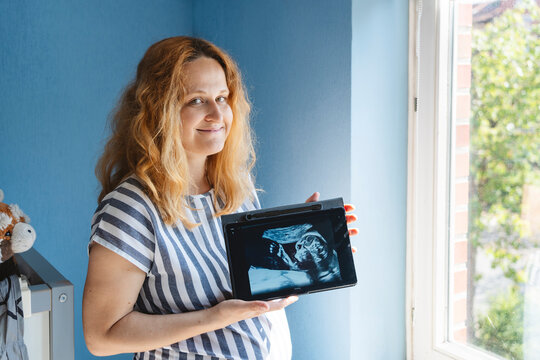 Happy pregnant woman showing ultrasound picture on tablet PC at home