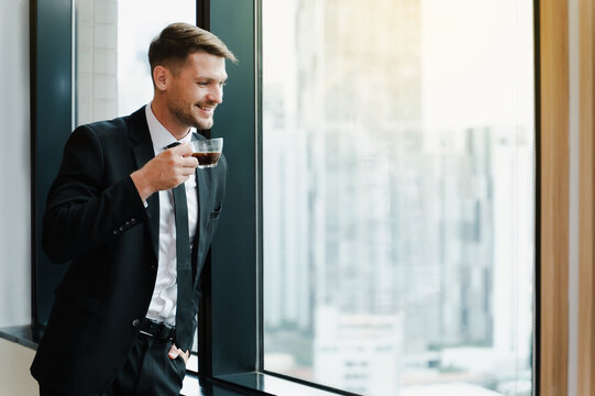 Businessman holding drinking coffee cup in office while looking through window,having break starting day in office.