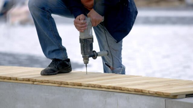 Employee uses drill to create holes in bench boards in park. Worker fastens wooden planks together and forms bench during renovation works