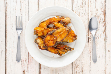 tasty slice fried nile tilapia fish in white circle ceramic plate with fork and spoon on white wood texture background, top table view, flat lay