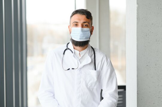 Male Pensive Thoughtful Arab Man Therapist Surgeon Wear White Uniform Medical Face Mask Standing At Workplace Looking Out Window Preventing Spread Infection Pandemic Outbreak Close-up Doctor Portrait.