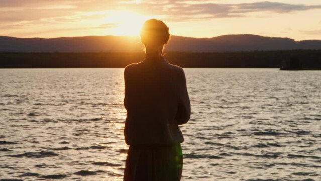 A lonely girl timidly stands on the shore of the lake at sunset and admires the beauty of nature. Harmony of nature and man.