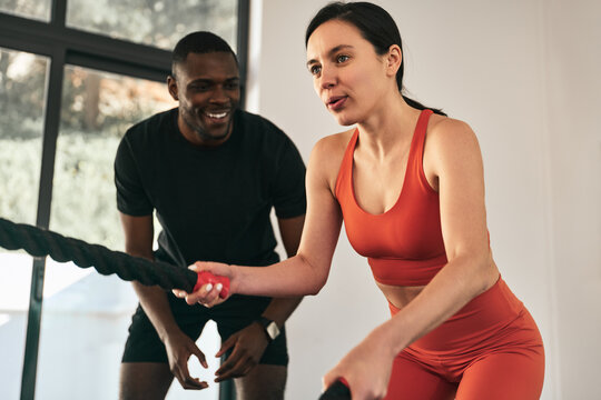 Smiling black trainer helping sportswoman doing exercise with battle ropes