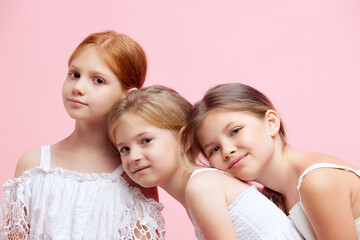 Three beautiful, cute little girls, children in ponytails, in white clothes looking at camera against pink studio background. Concept of skincare, childhood, cosmetology, health, beauty, wellness, ad