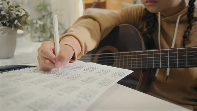 Teenage student making notes in copybook while learning to play guitar sitting at home
