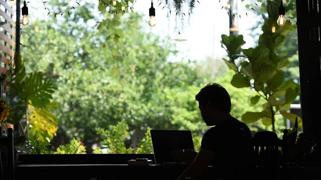 silhouette of man working on laptop smart phone and book sitting front of window green garden cafe coffee with passing traffic people and car