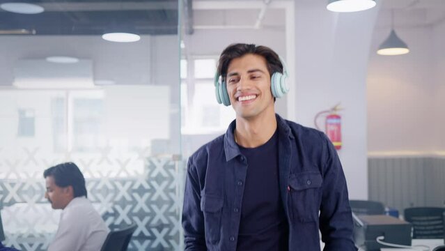 Young happy modern Indian Asian hipster male or man wearing headphones  listening to music and greeting colleagues or employees with smile while walking through the start up creative office workplace.