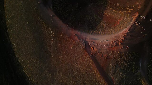 Aerial landscape view of tourists walking on an ancient inactive crater of volcanic magma rock on the Etna volcano mount in Sicily with a view of the city of Catania at sunset lights.