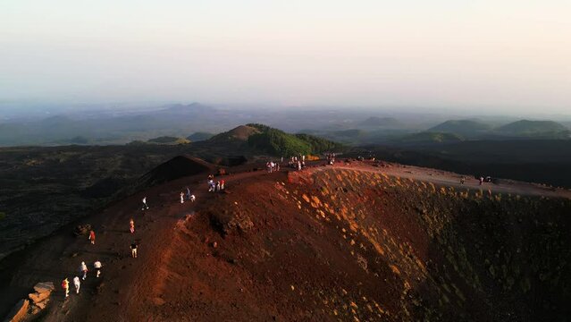 Aerial landscape view of tourists walking on an ancient inactive crater of volcanic magma rock on the Etna volcano mount in Sicily with a view of the city of Catania at sunset lights.
