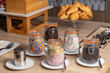 Fototapeta na wymiar hotel breakfast buffet. a wooden table topped with jars filled with food and sprinkles and croissants in the background