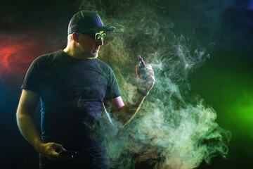 Man smokes vape. Guy with electronic cigarette. Man smoker uses vaping. Male is holding ecig device. Hipster with vape gadget in neon green light. Use of vape technologies. Alternative to nicotine - obrazy, fototapety, plakaty
