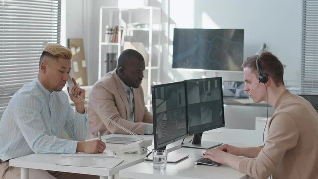 Multiracial male security service team watching outdoor surveillance cameras on computer monitors and making phone calls while working in modern office at daytime