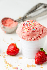 Top view of strawberry ice cream with colorful nuggets in white bowl on white table with strawberries, crumbles and serving spoon, selective focus, vertical, with copy space