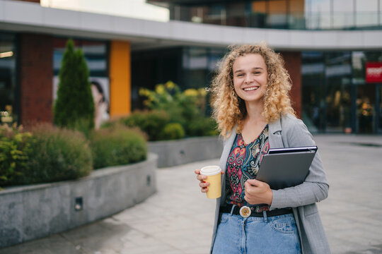 Curly-haired woman with takeaway caffeine beverage and closed laptop computer in urban city. Talking, working together, smiling, stylish freelance people