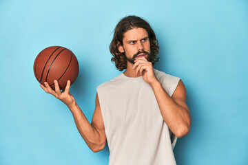 Bearded man with basketball in blue studio looking sideways with doubtful and skeptical expression.