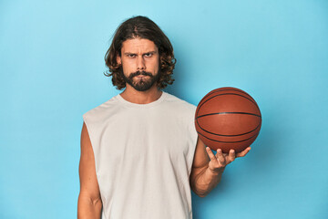 Bearded man with basketball in blue studio confused, feels doubtful and unsure.
