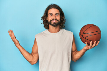 Bearded man with basketball in blue studio receiving a pleasant surprise, excited and raising hands.