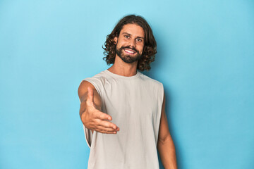 Bearded man in a tank top, blue backdrop stretching hand at camera in greeting gesture.