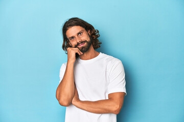 Bearded man in a white shirt, blue backdrop smiling happy and confident, touching chin with hand.