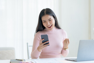 Happy young Asian woman raising hands with victory smiling happily with smart phone at home. Success, win, victory, triumph, congratulation, concept.