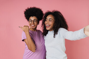 Two african women taking selfie and pointing away at copy space while standing isolated over pink background