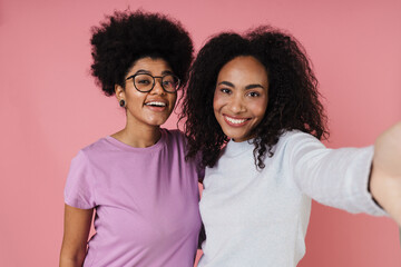 Two cheerful african women taking selfie while standing isolated over pink background