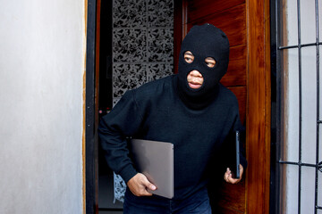 Sneaky and scared intruder wearing black balaclava mask stealing laptop from house. House intruder, housebreaking and burglary concept - obrazy, fototapety, plakaty