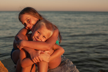 Brother and sister are sitting on the rocks on the seashore, at sunset. hugging and looking at the camera