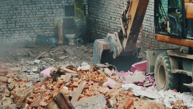View of bucket excavator raking up pink rubbish and concrete debris, turning, dumping garbage into truck. Clearing the destroyed building from remains of the foundation