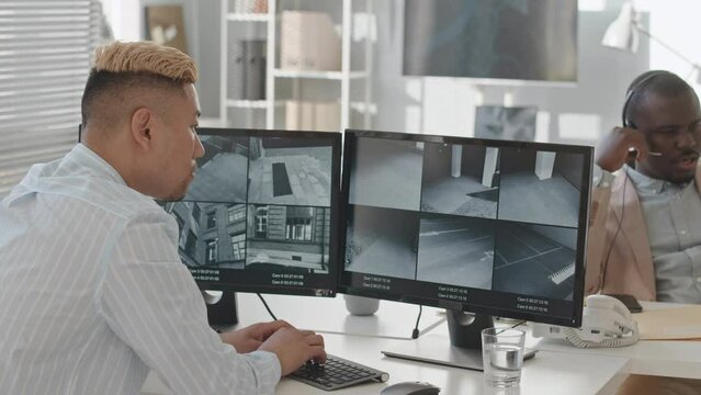 Back view slowmo of male Asian security service supervisor watching outdoor CCTV cameras on computer monitor while working in modern office with African American colleague