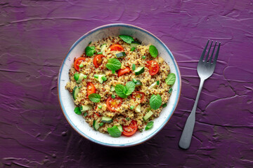 Quinoa tabbouleh salad in a bowl, a healthy dinner with tomatoes and mint, overhead shot with a fork on a purple background