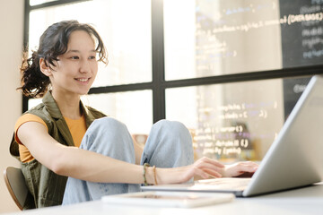 Smiling student sitting at desk and working online on laptop in the classroom