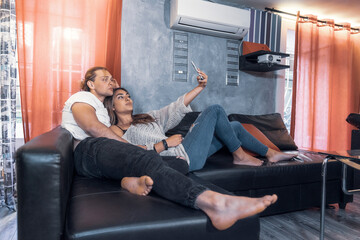 Cheerful couple of romantic caucasian guys smile taking a selfie at night in living room messaging and watching their cellphone device. Male fun guy enjoyng life looking a smart phone with a girl