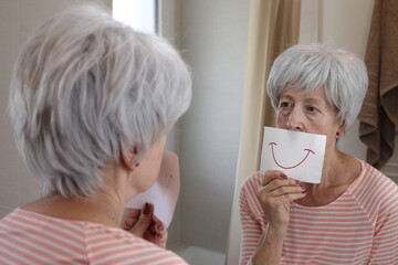 A senior woman with short gray hairstyle is holding a drawing with a smile in front of the mirror
