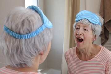 A sleepy looking senior woman in her sixties is staying in front of the mirror - obrazy, fototapety, plakaty