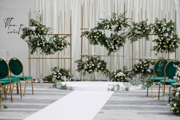 A luxurious wedding ceremony area. White flowers and green plants on golden stands. Emerald chairs with golden legs.